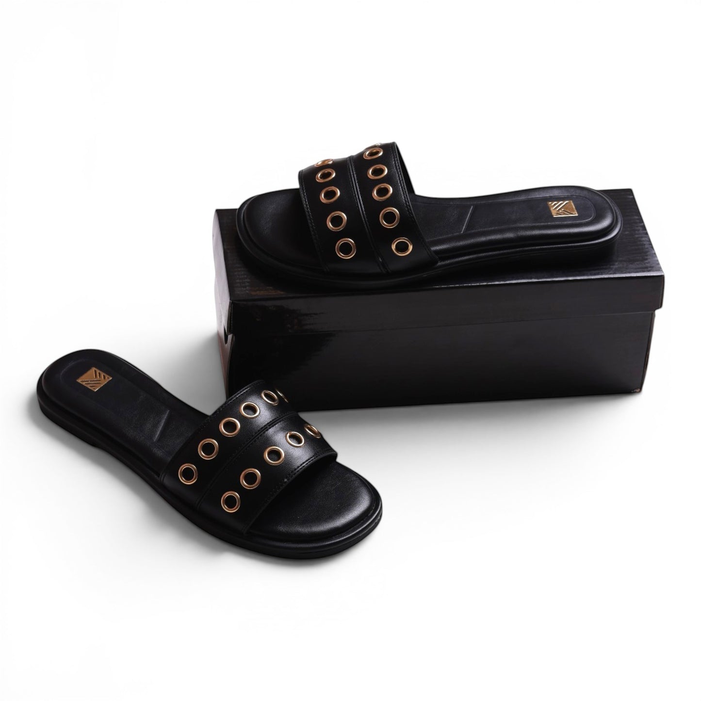 Moda Ug black leather slide sandals with double straps featuring gold eyelets. Flat sole, open toe.