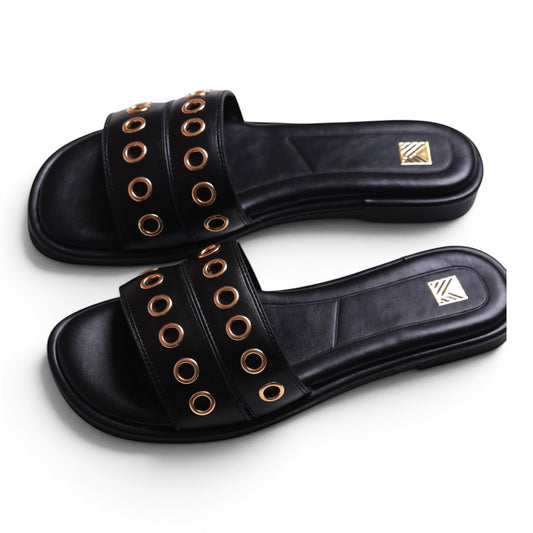 Moda Ug black leather slide sandals with gold eyelet details and a flat sole.