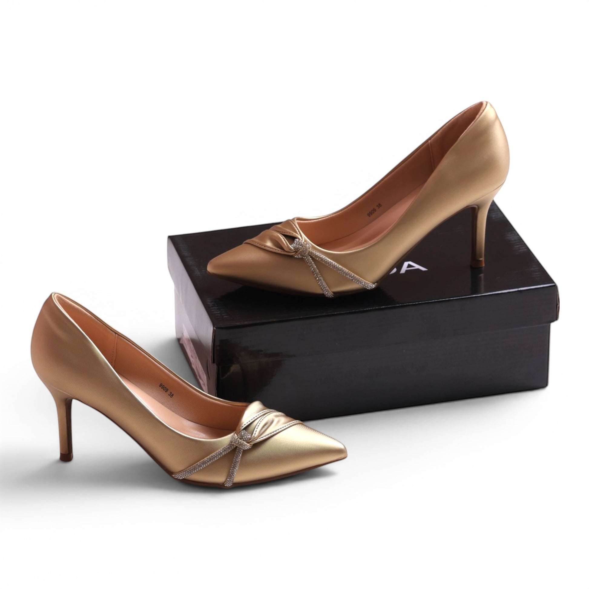 Moda Ug gold pointed-toe pumps with a knot and rhinestone detail on a black box.